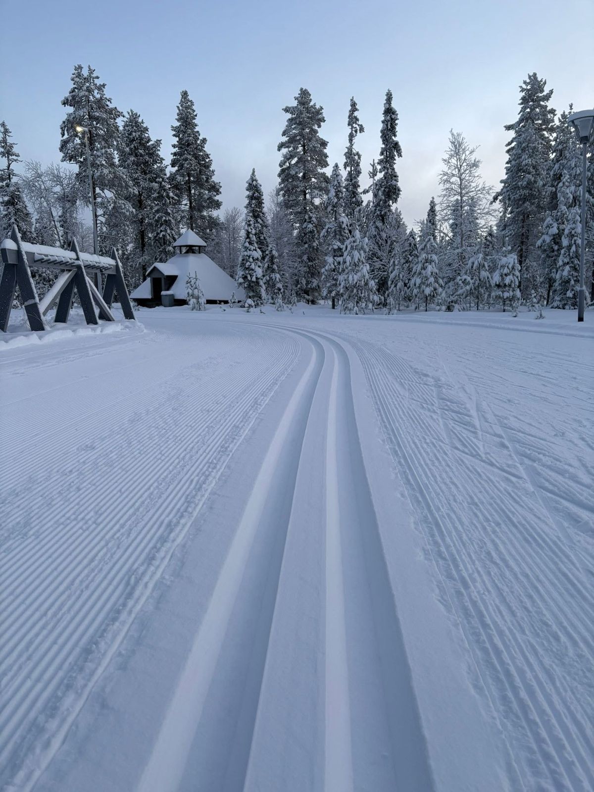 Vastavedetty latu kaartuu lumisten kuusten keskellä, taustalla kota ja suksiteline.